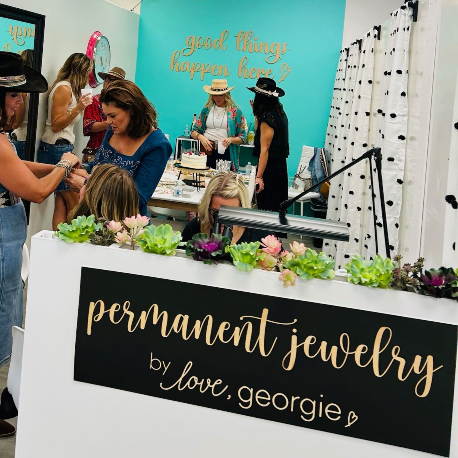 Event or trade show booth with 'permanent jewelry by love, georgie' sign and people around.