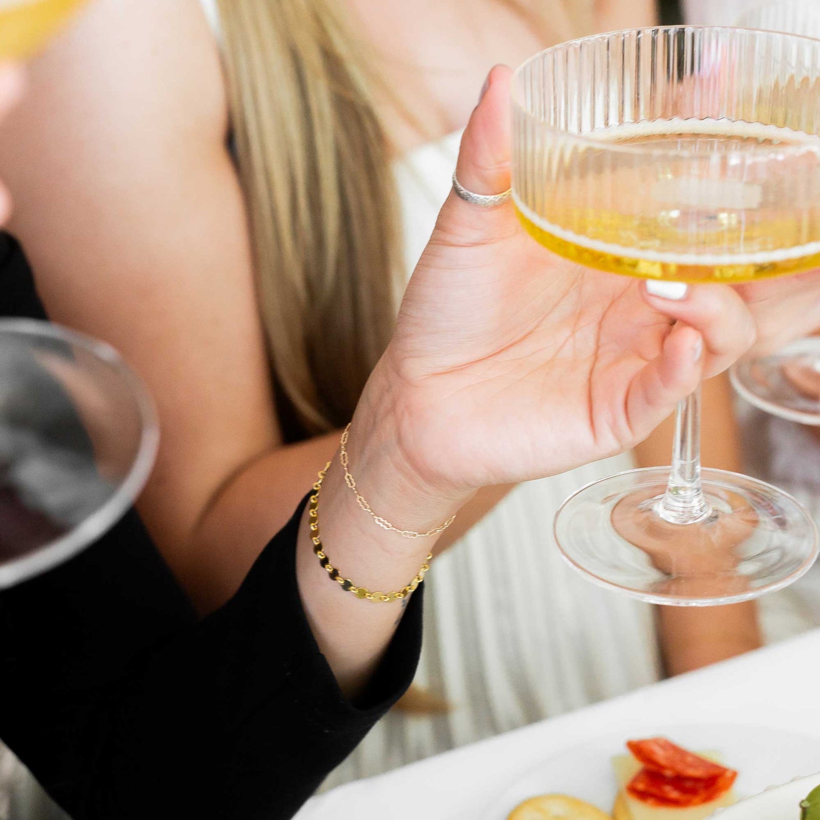 Person holding a glass up displaying her two gold bracelets with a blurred background