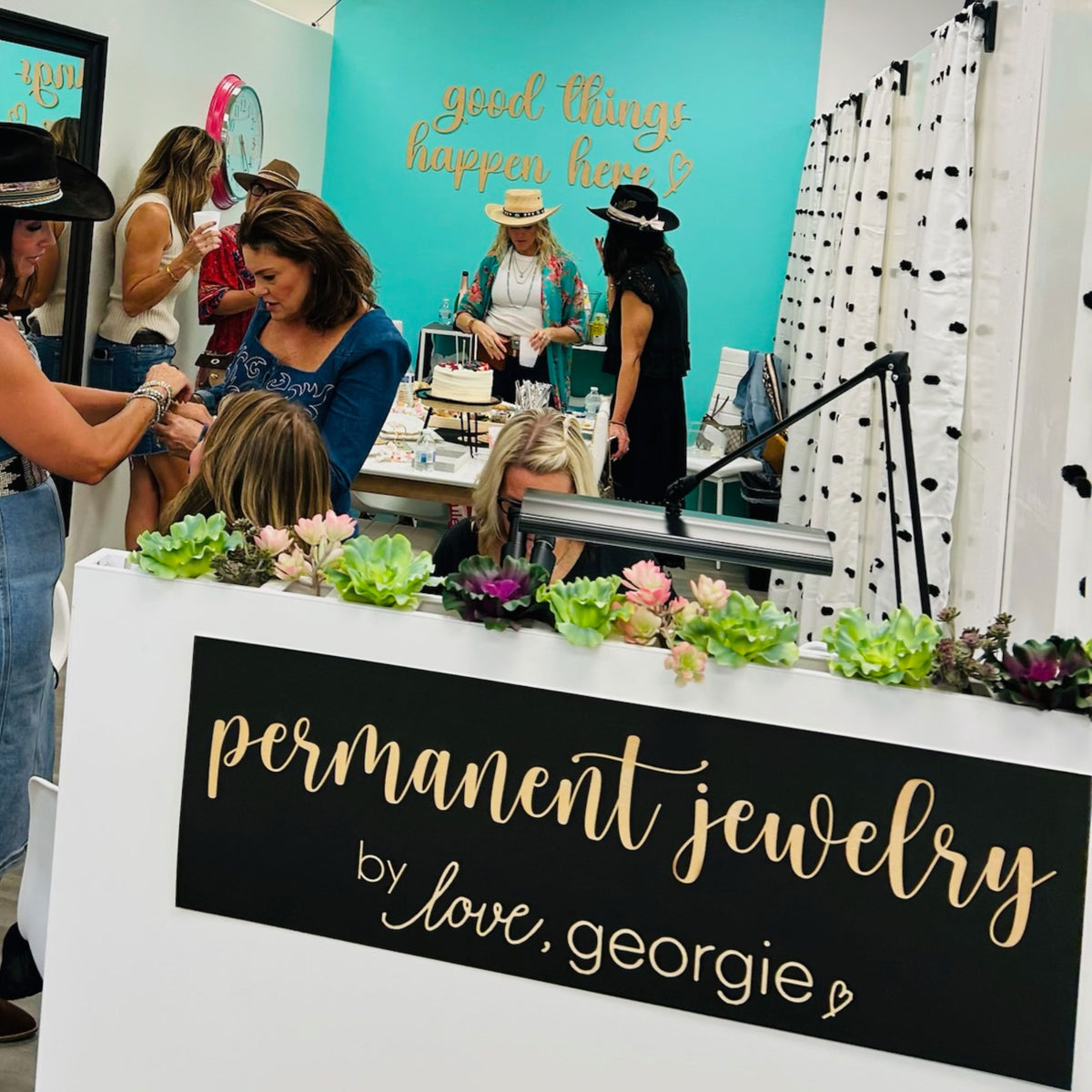 Event or trade show booth with 'permanent jewelry by love, georgie' sign and people around.