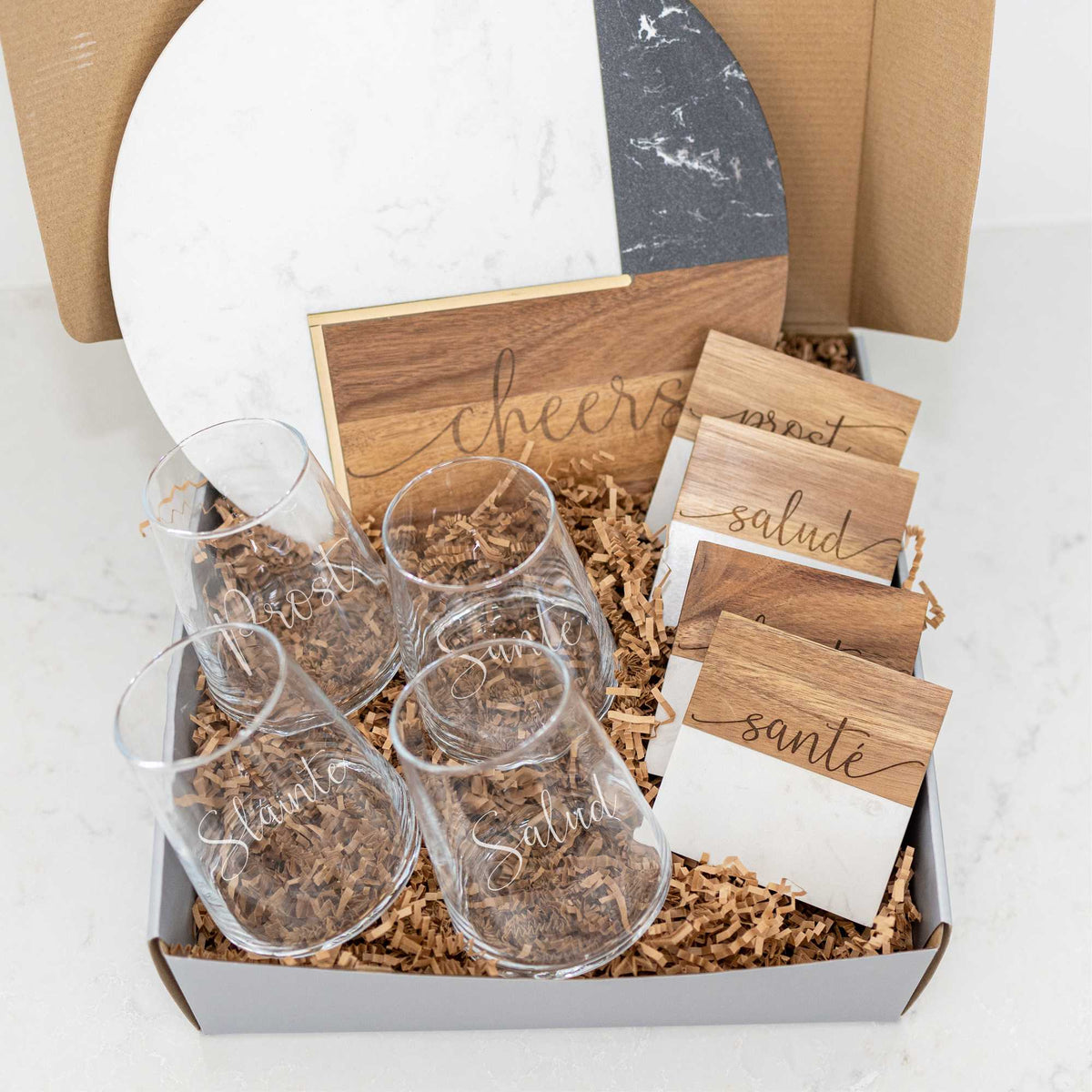 a fit box filled with four stemless wine glasses, four marble and wood coasters, and a round marble and wood charcuterie board. all items are engraved with international terms for &quot;cheers&quot;. 