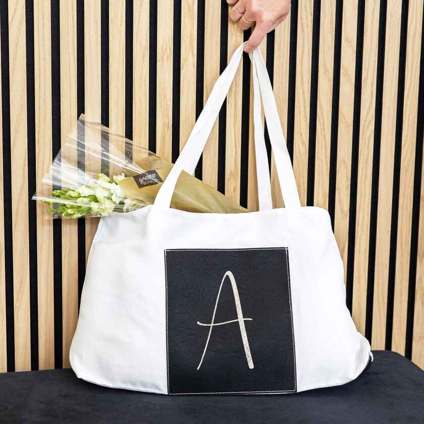 a white canvas tote bag with a black vegan leather pocket on outside is customized with a large letter "A" on it. tote bag is full and has a flower bouquet in it. 