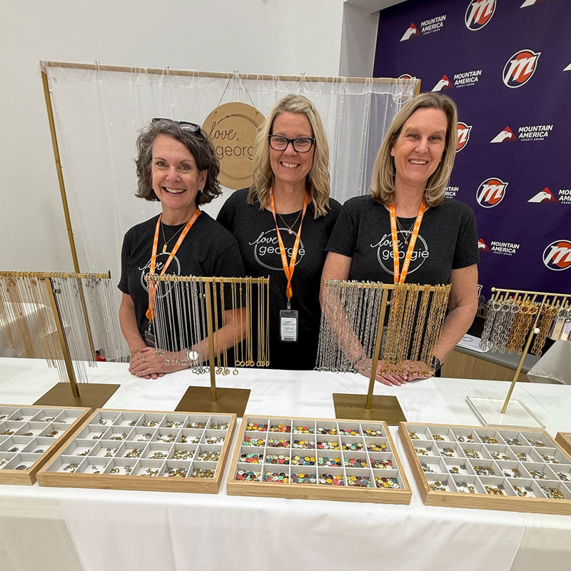 Three women standing behind a table with jewelry displays at an event.