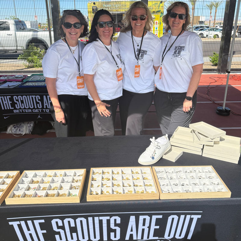 Four women in matching white shirts with 'The Scouts Are Out' sign and display of charms on a table.