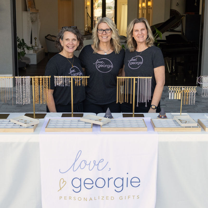 Three women in matching black shirts with 'love georgie' branding stand behind a display of jewelry.