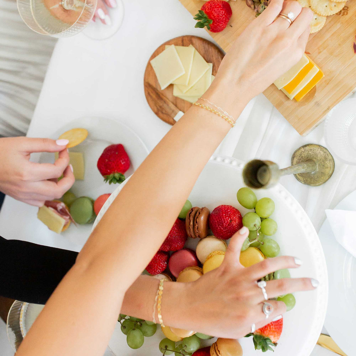 Two people picking fruits and cheese off a plate and a wooden serving board with a white tablecloth.