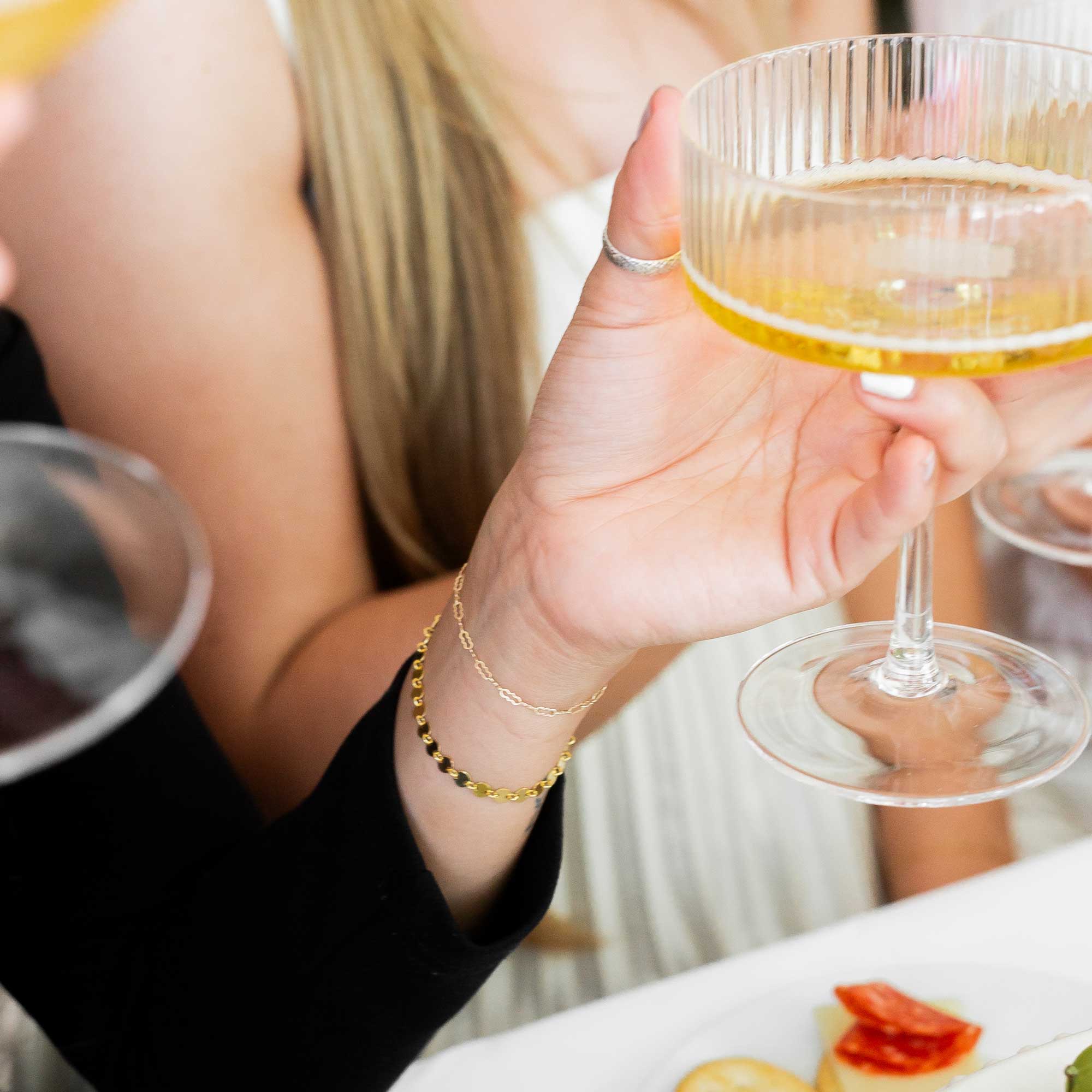 Person holding a glass up displaying her two gold bracelets with a blurred background