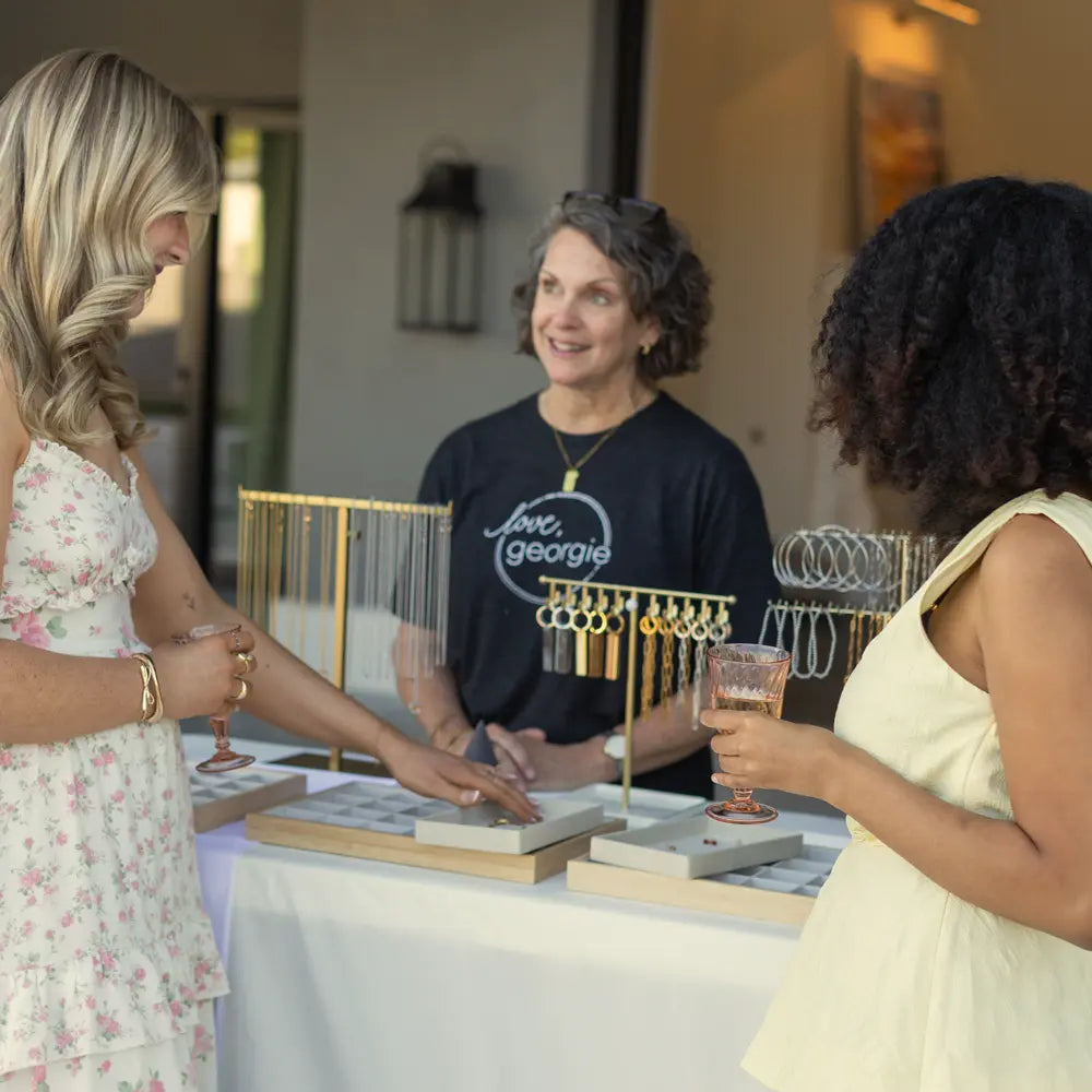 Three women interacting around a charm bar table with jewelry displays.