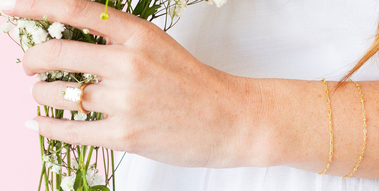 Hand holding a small bouquet of flowers with a gold ring and bracelet on a light background
